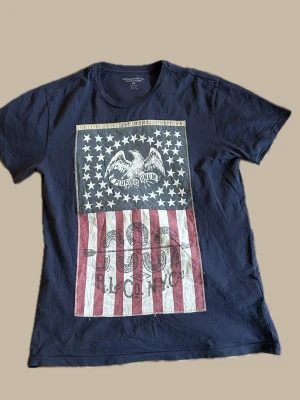 Mörkblå Ralph Lauren t-shirt med amerikanskt tryck - Säljer en mörkblå t-shirt från Ralph Lauren Denim & Supply med stort amerikanskt flaggtryck framtill. Trycket har stjärnor, örn och texten 'E Pluribus Unum' samt röda och vita ränder. Klassisk passform och rund hals. Perfekt för dig som gillar statement-plagg.