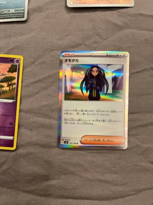 Japanskt Pokémonkort Omotaka holo - Säljer ett japanskt Pokémonkort med motivet Omotaka. Kortet är holografiskt med färger i blått, lila och orange. Texten är på japanska och kortet har en klassisk rektangulär form. Perfekt för samlare av Pokémonkort eller dig som gillar unika kort.