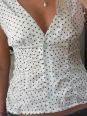 Polka dot topp  - Söt vit blus med svarta prickar och djup v-ringning. Blusen har knappar framtill och figurnära passform som framhäver midjan. Perfekt för dig som gillar en feminin och trendig stil. Passar s-xs