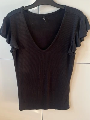 Svart ribbad topp från ONLY - Snygg svart ribbad topp från ONLY med v-ringning och volangdetaljer på ärmarna. Toppen har en figurnära passform och är tillverkad i mjuk stretchig bomullsblandning. Perfekt att styla med jeans eller kjol för en trendig look.