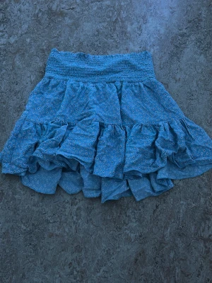 Blå volangkjol med småblommigt mönster - Supersöt blå kortkjol med volanger och små vita blommor. Kjolen har bred smock i midjan för skön passform och ett luftigt, flowy tyg som passar perfekt till varmare dagar. Perfekt för dig som vill ha en lekfull och trendig look.