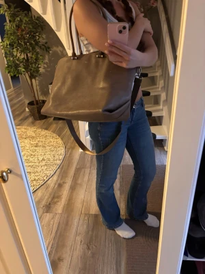 Brun axelremsväska i skinn - Säljer en stilren brun handväska i slätt skinn med både handtag och justerbar axelrem. Väskan har en enkel, fyrkantig form och stängs upptill. Perfekt för dig som gillar minimalistisk design och vill ha plats för allt du behöver.