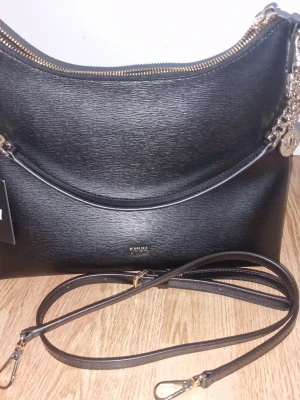 Svart handväska från DKNY - Snygg svart handväska från DKNY i strukturerat läder med guldfärgade detaljer och kedja. Väskan har en avtagbar axelrem och dragkedja upptill. Medföljer original dustbag med DKNY-logga.