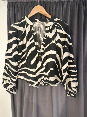 Zebramönstrad blus med knappar - Snygg blus med svartvitt zebramönster och knappar framtill. Blusen har lång ärm med resår vid ärmslut och en avslappnad passform. Perfekt för dig som vill sticka ut med ett djuriskt mönster.