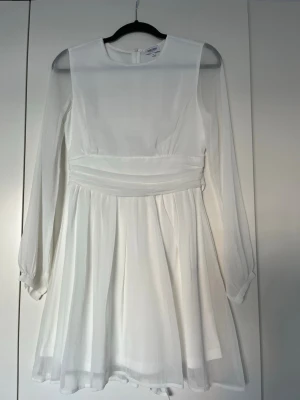 Vit kort klänning med knytband - Säljer en elegant vit kortklänning med långärmade transparenta ärmar och rund halsringning. Klänningen har ett markerat midjeband och knytband i ryggen för extra detalj. Tillverkad i ett lätt, flödande material som ger en luftig känsla.