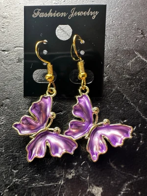 Lila fjärilsörhängen i metall - Unika örhängen i form av fjärilar med skimrande lila vingar och guldfärgade detaljer. Hängande modell med krok, perfekta för dig som vill sticka ut. Tillverkade i metall med emaljerad yta för extra glans.