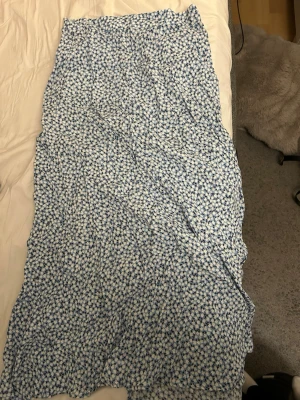 Blommig blå långkjol med slits - Säljer en långkjol med blått och vitt blommönster. Kjolen har en rak passform och en snygg slits framtill.