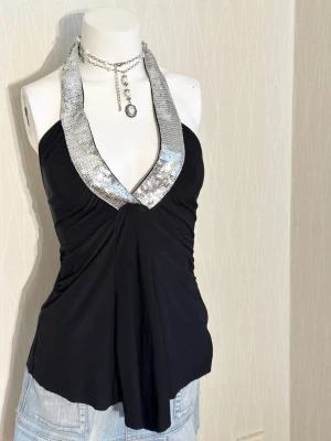 Svart halterneck topp med paljetter - Snygg svart halterneck topp med bred, glittrig silverkant dekorerad med paljetter. Djup v-ringning och lätt utsvängd passform. Perfekt för dig som vill sticka ut och glänsa lite extra.