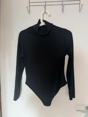 Svart ribbad body med polokrage Boohoo M eu 40 uk 12 - Snygg svart body med lång ärm och polokrage. Tillverkad i ribbat stretchigt material som sitter tajt och bekvämt. Perfekt att styla med jeans eller kjol för en trendig look.