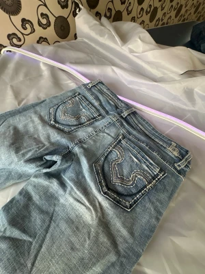 Blå bootcut jeans med kontrastsömmar - Säljer ett par ljusblå bootcut jeans med slitningar och tydliga kontrastsömmar på bakfickorna. Jeansen har klassisk femficksmodell och låg midja. Perfekta för en avslappnad och trendig look.