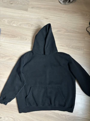 Svart Essentials Fear of God hoodie - Säljer en svart hoodie från Essentials Fear of God med stor ficka framtill och huva. På ryggen står det 'ESSENTIALS FEAR OF GOD' i svart text. Hoodien har ribbade muddar vid ärmslut och nederkant. Perfekt för en avslappnad och trendig streetwear-look.