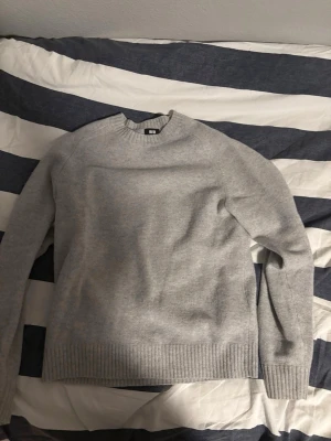 Ljusgrå stickad tröja Uniqlo - En stilren ljusgrå stickad tröja från Uniqlo med rund halsringning och ribbade muddar vid ärmslut och nederkant. Perfekt basplagg som är lätt att matcha och har en mjuk, skön känsla. Passar dig som gillar enkel och clean stil.