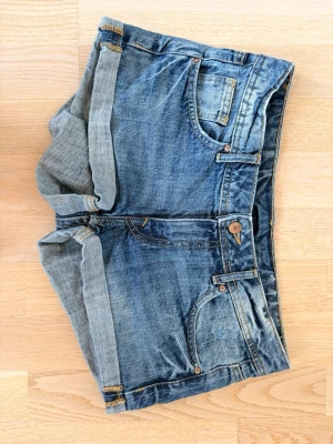 Blå jeansshorts med uppvik - Snygga blå jeansshorts med uppvikta benslut och klassiska fem fickor. Shortsen har en knappgylf och bälteshällor. Perfekta för varma dagar och ger en avslappnad look.