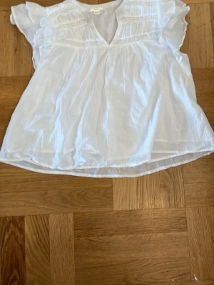 Vit blus med volangärmar och spetsdetaljer - Söt vit blus med volangärmar och v-ringning. Blusen har fina spetsdetaljer framtill och är gjord i ett tunt, luftigt tyg. 