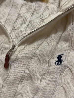 Vit kabelstickad tröja Polo Ralph Lauren - Vit kabelstickad tröja från Polo Ralph Lauren med halv dragkedja och blå broderad logga på bröstet. Tröjan har hög krage och ribbstickade detaljer. Perfekt för dig som gillar klassisk och stilren design.