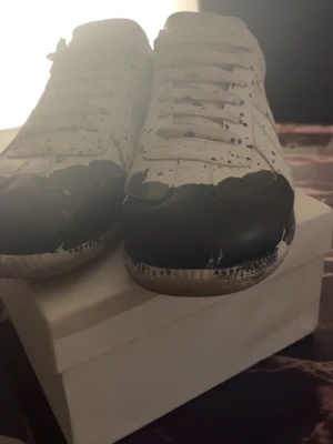 Svarta och vita maison margiela skor  - Unika sneakers i svart och vitt med coola färgstänk och slitna detaljer. Skorna har klassisk låg profil, vita snören och svart tåparti. Perfekta för dig som vill sticka ut med din stil.
