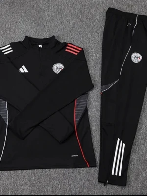 Svart träningsset från Adidas - Säljer ett svart träningsset från Adidas med långärmad tröja och byxor. Setet har vita och röda detaljer, dragkedja vid halsen och klubbmärke på både tröja och byxa. Tillverkat i funktionsmaterial som andas, perfekt för träning eller uppvärmning.
