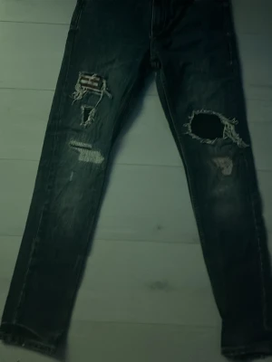 Mörkblå slitna jeans med hål - Säljer ett par mörkblå jeans med tydliga slitningar och stora hål på båda benen. Jeansen har raka ben och en klassisk femficksmodell. Perfekta för dig som gillar en edgy och avslappnad stil.