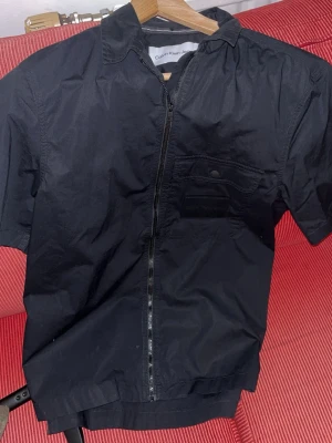 Svart overshirt  från Calvin Klein  - Snygg svart jumpsuit från Calvin Klein Jeans med korta ärmar, dragkedja framtill och bröstficka med knapp. Klassisk krage och enkel, stilren design som passar till många tillfällen. Perfekt för dig som gillar en clean och modern look.