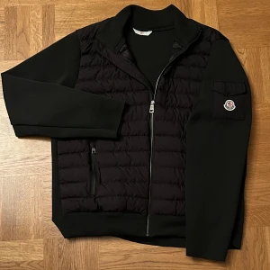 Svart cardigan från Moncler - Svart cardigan från Moncler med quiltad front, släta ärmar och Moncler-logga på ärmficka. Jackan har dragkedja framtill, två sidofickor med dragkedja och innerfoder med serietidningsdetalj. Perfekt för dig som gillar stilren och sportig look.