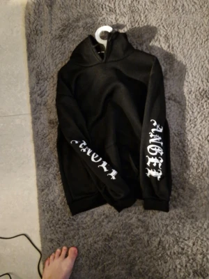 Svart hoodie med vita tryck och vingar - Säljer en svart hoodie med huva och feta vita tryck på ärmarna där det står 'ANGEL' och 'CHILD'. På ryggen finns ett stort tryck med vita änglavingar. Hoodien har en avslappnad passform och är gjord i mjukt material, perfekt för en cool streetwear-look.