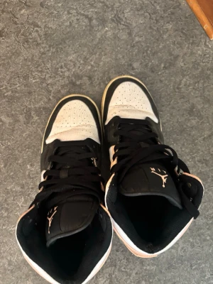 Air Jordan 1 Mid svart/rosa/vit - Snygga Air Jordan 1 Mid sneakers i svart, rosa och vitt. Skorna har klassisk hög siluett, svart snörning och ikoniska loggor på plösen och sidan. Ovandelen är i läder och textil, med perforerad tåbox för extra ventilation. Perfekta för dig som gillar streetwear.