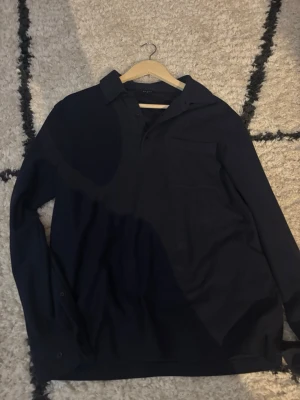 Mörkblå långärmad skjorta - Snygg mörkblå långärmad skjorta med klassisk krage och bröstficka. Skjortan har knappar vid ärmsluten och är tillverkad i ett mjukt tyg som känns bekvämt mot huden. Perfekt för dig som gillar stilrena och enkla plagg.