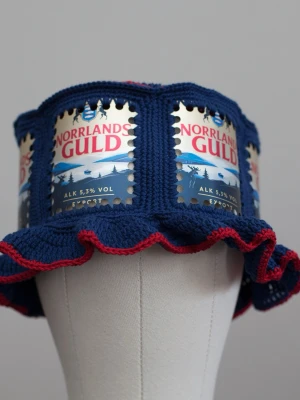 Virkad ölhatt Norrlands Guld öl - Unik virkad ölhatt med Norrlands Guld.  Virkad i 100% bomull. Passar upp till ca 60cm.  Vissa burkar kan ha mindre repor eller bucklor, vilket tyvärr kan uppstå under tillverkningen av hattarna. Insidan av burkarna slipas efter hålen tagits för att undvika vassa kanter. Därför kan det ibland finnas lite slipmärken, men det är inget som stör eftersom det är på insidan av hattarna.  Färger kan variera något beroende på skärminställningar.