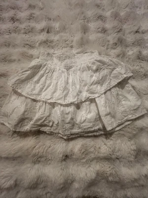 Vit volangkjol med broderi - Supersöt vit kjol med dubbla volanger och broderade blommor. Kjolen har bred resår i midjan för skön passform och är gjord i ett lätt bomullstyg. Perfekt för dig som gillar romantisk och feminin stil. Seriöst jätte fin på och helt perfekt till sommaren nu också.