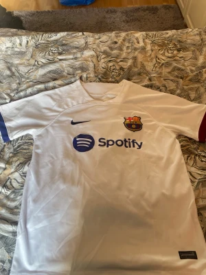 Vit FC Barcelona matchtröja Nike - Säljer en vit FC Barcelona matchtröja från Nike med klubbmärke och Spotify-logga på bröstet. Tröjan har korta ärmar med blå och vinröda detaljer, samt UNHCR-logga på ryggen. Perfekt för dig som älskar fotboll och vill stötta Barca.