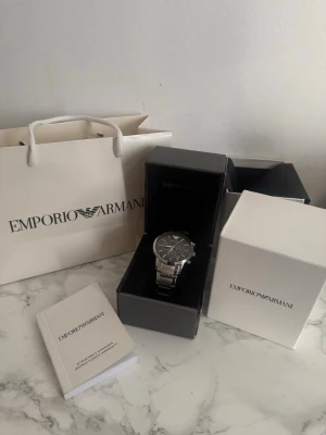 Emporio Armani  klocka svart  - Stilren klocka från Emporio Armani med silverfärgat armband i metall och svart urtavla. Klockan har kronograf-funktion, datumvisning och klassisk design med märkets logga vid tolvslaget. Perfekt accessoar för dig som gillar en clean och elegant look.