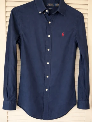 Marinblå skjorta från Polo Ralph Lauren - Klassisk marinblå skjorta från Polo Ralph Lauren med vit knappslå och röd broderad logga på bröstet. Skjortan har lång ärm, button-down krage och är tillverkad i bomull. Perfekt för en stilren och tidlös look.