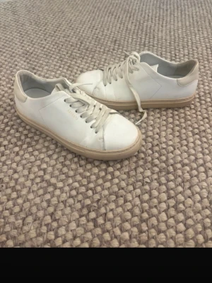 Vita sneakers från Axel Arigato - Stilrena vita sneakers från Axel Arigato i lågt snitt. Skorna har ovandel i slätt läder, vita skosnören och en beige sula. Diskret logga i guld på sidan. Perfekta för en clean och minimalistisk look.
