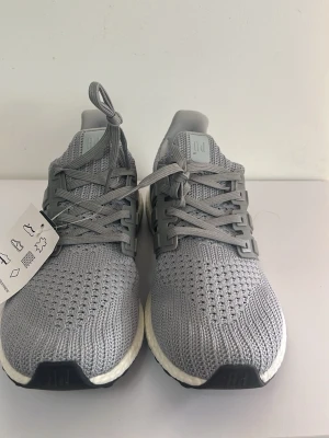 Grå Adidas Ultraboost sneakers - Snygga grå Adidas Ultraboost sneakers med stickad mesh-överdel och klassiska tre ränder på sidan. Skorna har en vit Boost-mellansula och svart yttersula, vilket ger både komfort och stil. Perfekta för dig som gillar sportig look och vill ha något trendigt.