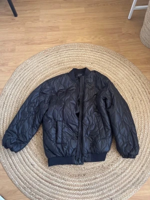 Svart quiltad bomberjacka Vero Moda - Snygg svart quiltad bomberjacka från Vero Moda med ribbade muddar vid ärmslut och nederkant. Jackan har dragkedja framtill och två sidofickor. Perfekt för dig som gillar en stilren och trendig look.