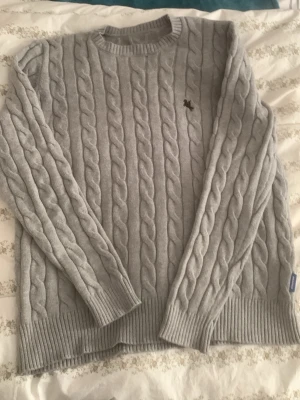 Ljusgrå kabelstickad tröja Lexington - Snygg ljusgrå kabelstickad tröja från Lexington med rund halsringning och ribbade muddar. Tröjan har ett diskret broderat logomärke på bröstet och är gjord i mjukt material som passar perfekt till kyliga dagar.