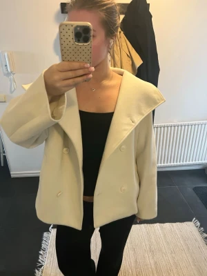 Krämvit kappa med bred krage - Snygg krämvit kappa med dubbelknäppning och bred, markerad krage. Modellen är rak  vilket ger en modern och stilren look. Perfekt att slänga över en outfit för en clean vibe. Materialet ser ut att vara ullblandning.