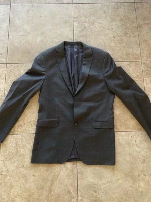 Mörkgrå klassisk kavaj - Snygg mörkgrå kavaj med två knappar framtill och två fickor med lock. Kavajen har en klassisk passform och är helfodrad med slät insida. Perfekt för en stilren look och passar till skjorta eller t-shirt. OBS! En knapp saknas tyvärr därav priset! 