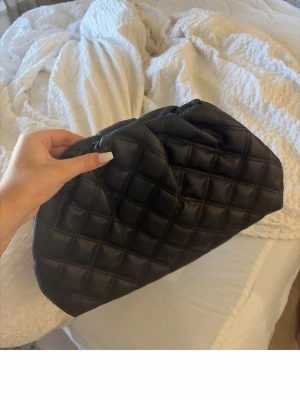 Svart clutch  - Säljer en svart clutch  i quiltad design med mjukt, vadderat material. Väskan har en puffig form och stängs upptill. Perfekt accessoar för att lyfta din outfit med en trendig touch.