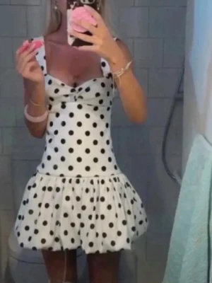 Vit kortklänning med svarta prickar - Supersöt vit kortklänning med svarta prickar och volangkjol. Klänningen har breda axelband och en liten rosettdetalj framtill. Perfekt för dig som vill sticka ut med en lekfull och trendig look.