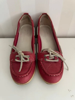 Röda seglarskor med vita snören - Säljer ett par klassiska röda seglarskor i läder med vita snören och kontrastsömmar. Skorna har en låg profil och vita detaljer längs sidorna. Perfekta för en avslappnad och stilren look.