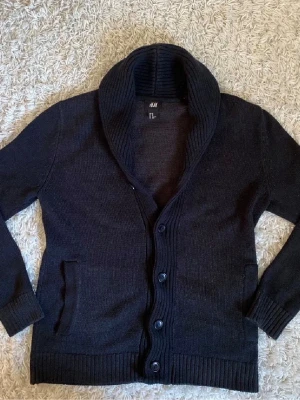 Svart kofta med sjalkrage från H&M - Mysig svart kofta från H&M med sjalkrage och knäppning framtill. Koftan har långa ärmar, ribbade muddar och två fickor fram. Perfekt för lager på lager och enkel att matcha med olika outfits.