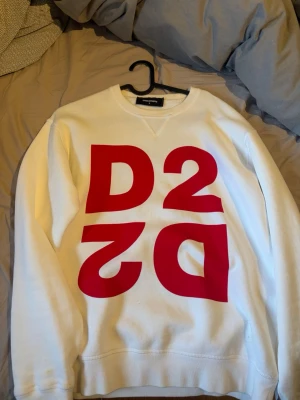 Vit sweatshirt med röd D2-logga - Cool vit sweatshirt från Dsquared2 med stor röd D2-logga på framsidan. Tröjan har rund halsringning och ribbade muddar vid ärmslut och nederkant. Perfekt för dig som gillar streetwear och vill sticka ut med en statement-piece.