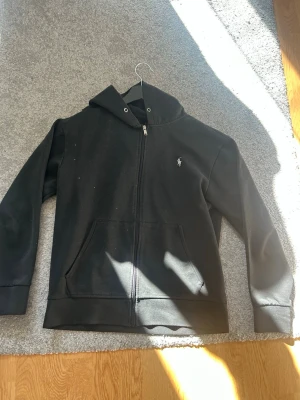 Svart hoodie från Polo Ralph Lauren - Klassisk svart hoodie från Polo Ralph Lauren med dragkedja framtill och vit broderad logga på bröstet. Hoodien har huva med dragsko och två stora fickor framtill. Tillverkad i mjuk bomullsblandning som känns skön mot huden.