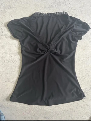 Svart topp med spetsdetalj - Snygg svart topp med korta ärmar och djup V-ringning dekorerad med spets. Figurnära passform och lätt glansigt material som ger en elegant look. .