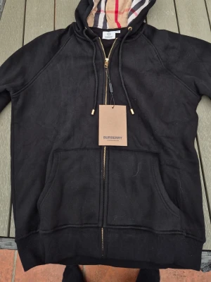 Svart hoodie från Burberry med dragkedja - Säljer en svart hoodie från Burberry med klassiskt rutigt foder i huvan och guldfärgade detaljer på dragkedja och snören. Hoodien har två stora fickor framtill och ribbade muddar. Perfekt för dig som gillar exklusiv streetwear och snygga detaljer.
