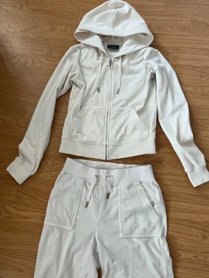 Vit velourset från Juicy Couture - Mysig vit loungewear-set från Juicy Couture i mjuk velour. Setet består av en zip-hoodie med fickor och dragsko samt matchande byxor med resårmidja och fickor. Perfekt för chill dagar hemma eller när du vill vara extra comfy.