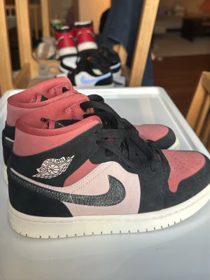 Air Jordan 1 Mid rosa och svart - Snygga Air Jordan 1 Mid sneakers i rosa, svart och vitt. Skorna har klassisk Jordan-logga på sidan, svart Swoosh och svarta skosnören. Ovandelen är i mocka och läder, med perforerad tåbox för extra ventilation. Perfekta för dig som vill sticka ut med stil.