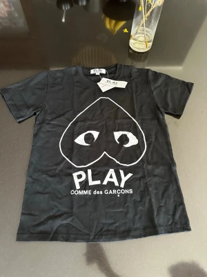 Svart Comme des Garçons PLAY t-shirt - Svart t-shirt från Comme des Garçons PLAY med det ikoniska hjärtmotivet och stora vita trycket framtill. Klassisk rund hals och korta ärmar. Tillverkad i mjuk bomull för en skön och avslappnad stil.