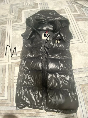 Svart glansig dunväst med huva - Snygg svart dunväst med glansig finish och huva. Västen har dragkedja framtill och broderad NY-logga på bröstet. Perfekt för lager-på-lager och streetwear-stil.